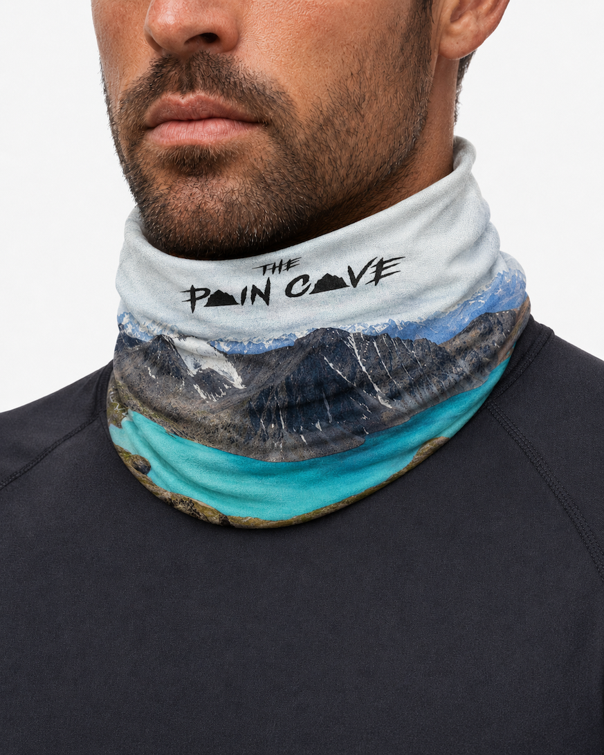 man wearing a buff/snood with a stunning view of large mountains with snow and a large lake.